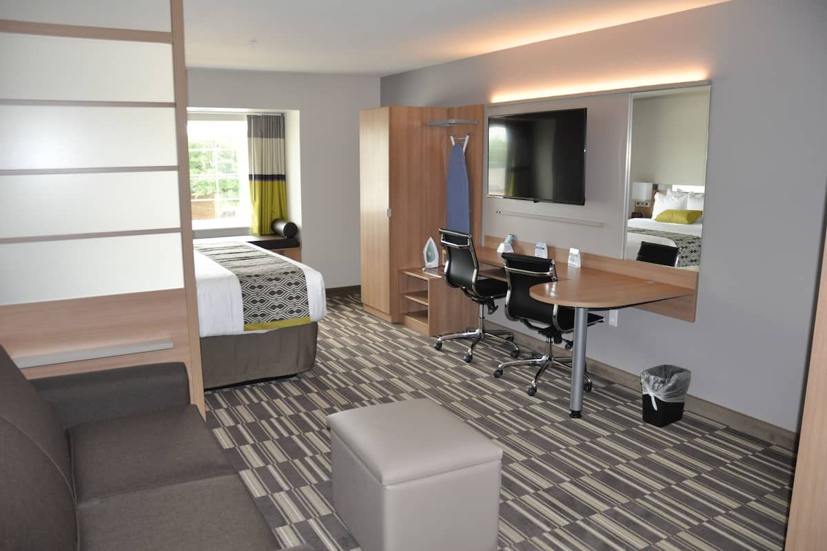A modern hotel room with a bed, a desk, a chair, a small table, a trash bin, and a large mirror.