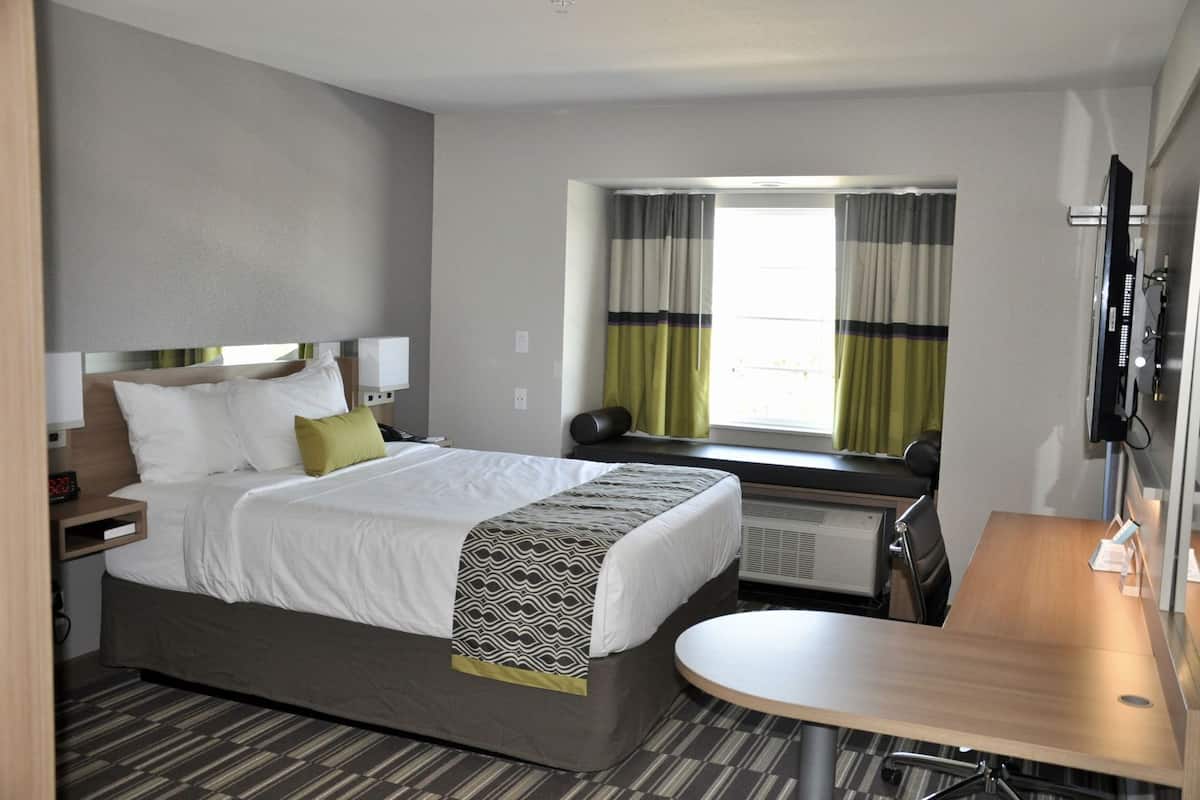 A hotel room with a bed, bedside table, desk, chair, window with curtains, and a television.