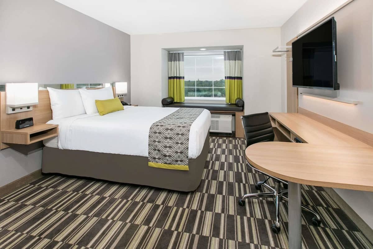 A hotel room with a bed, bedside table, TV, desk, chair, and a window with curtains.