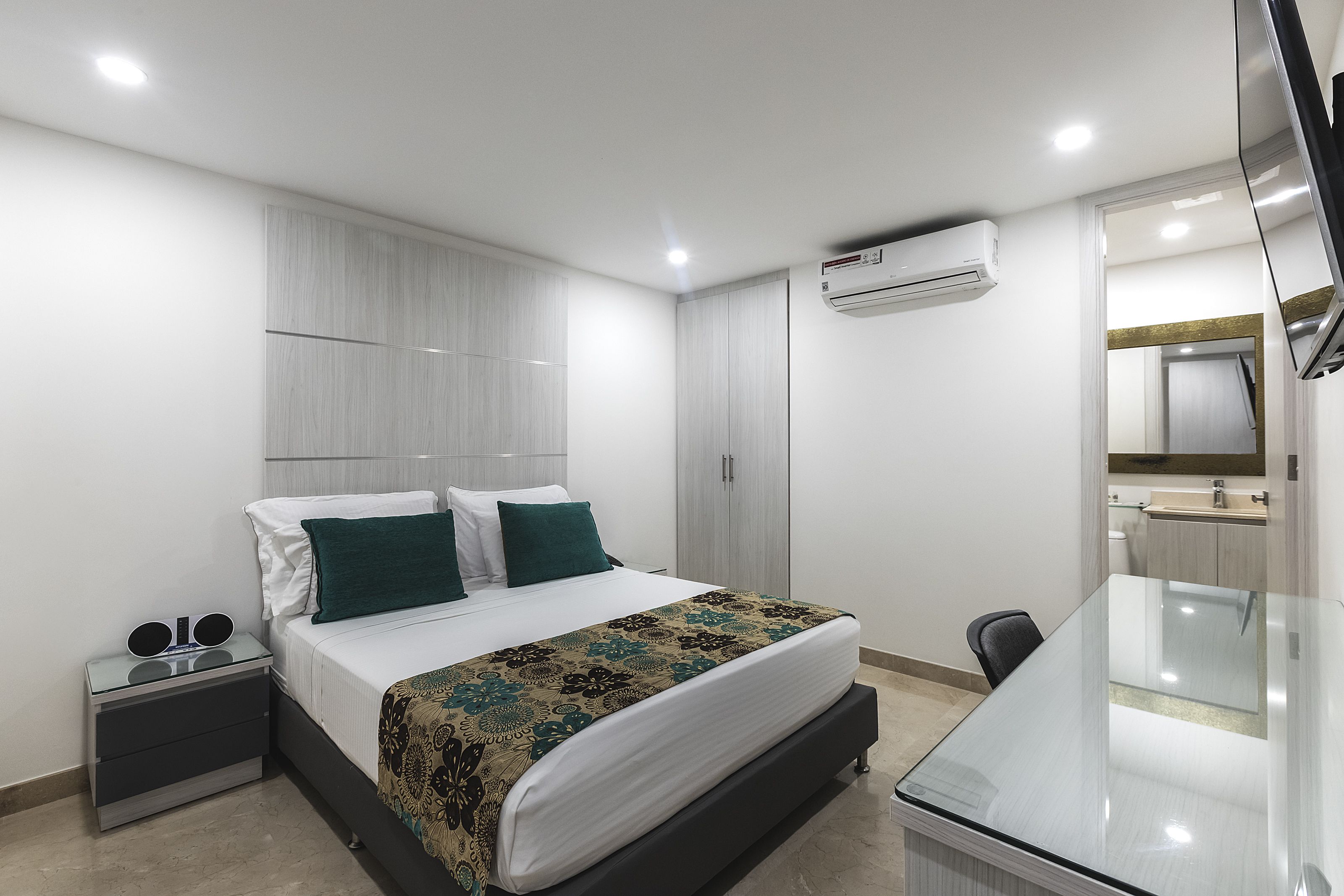 A modern hotel room with a large bed, a bedside table with a mirror, a desk with a chair, and an air conditioning unit on the wall.