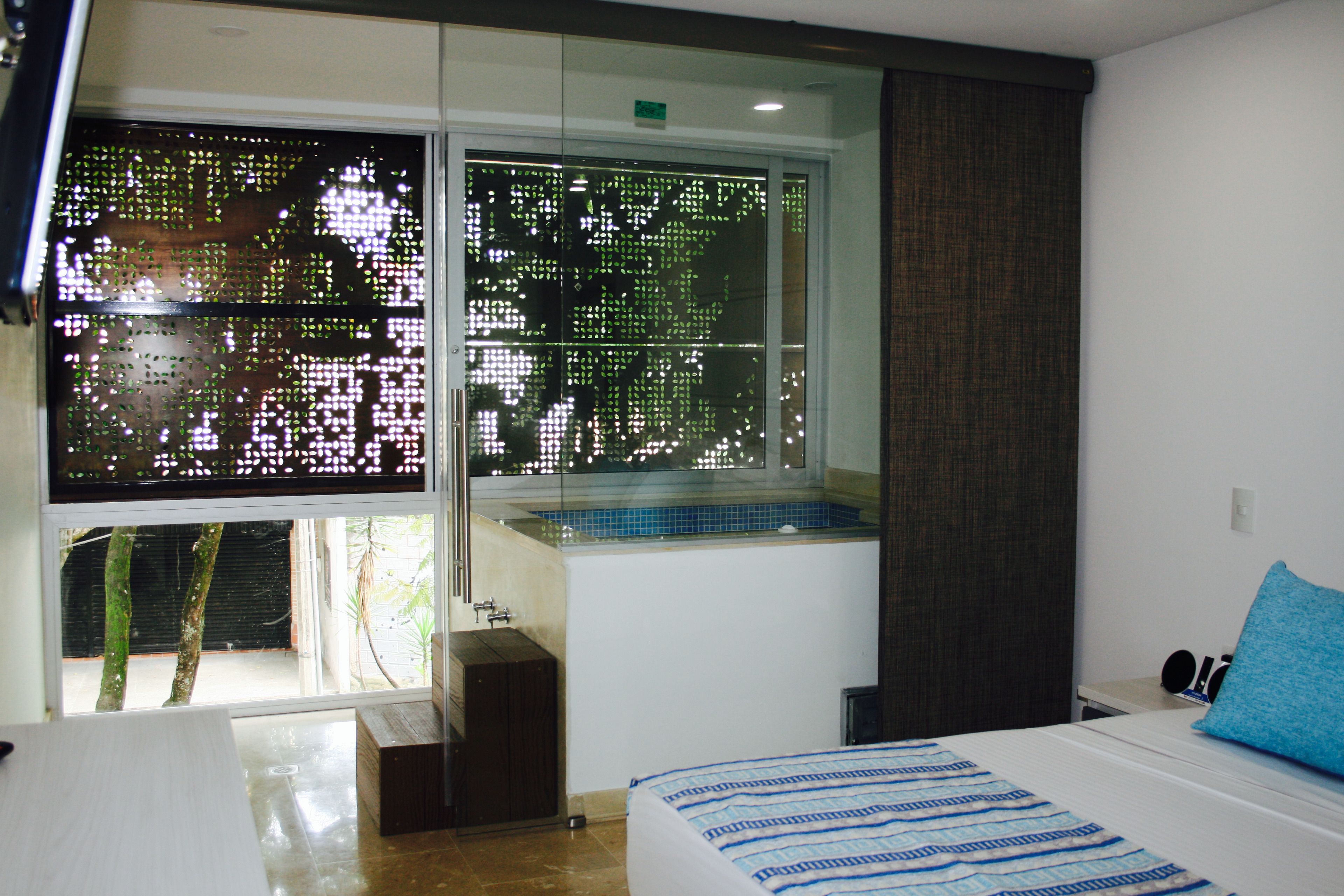 A modern hotel room with a large window, a bed with a striped bedspread, a small bedside table, and a blue pillow.