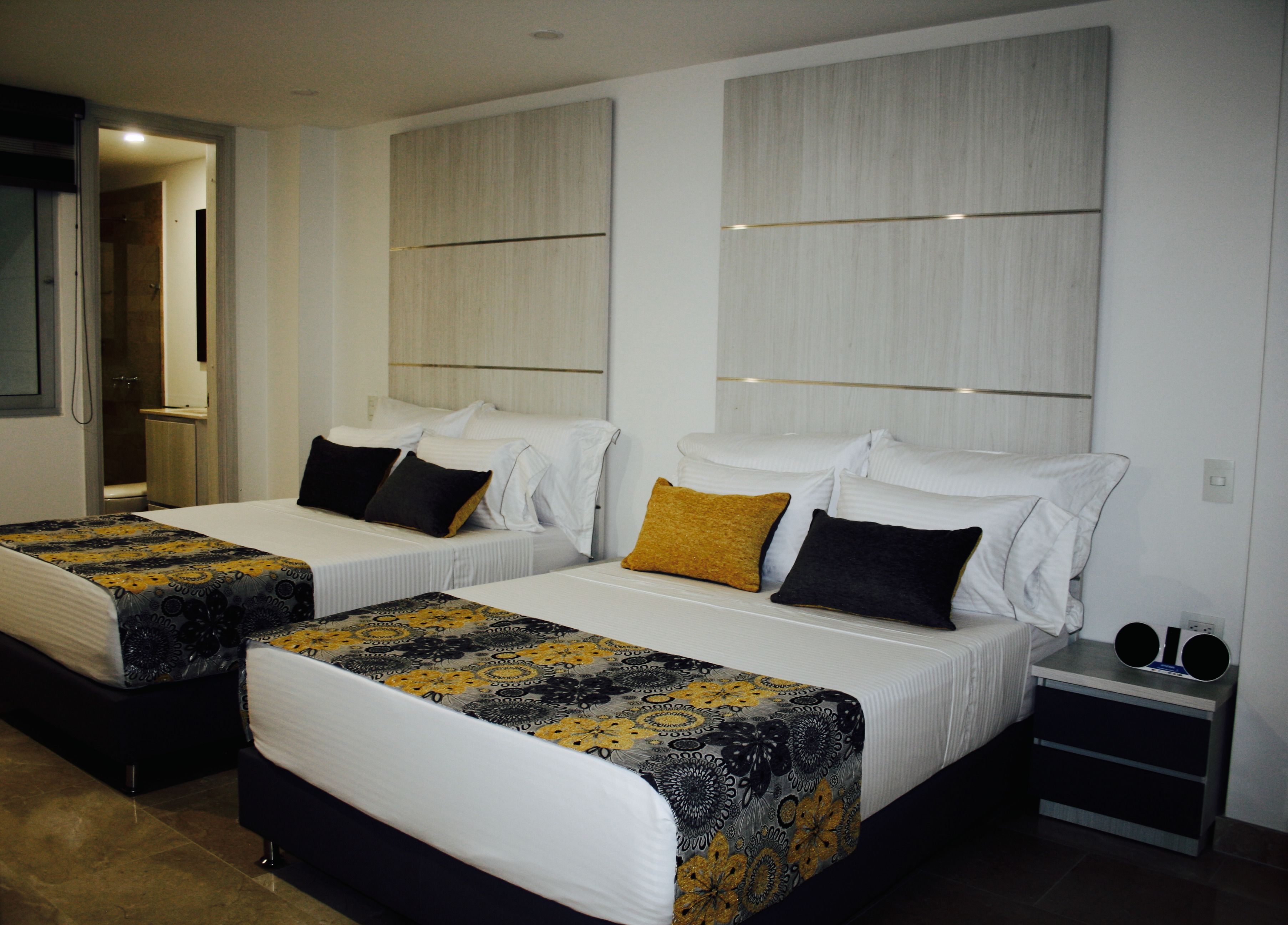 Habitación de hotel con dos camas, cada una con una colcha estampada, almohadas blancas y una mesita de noche con un altavoz.