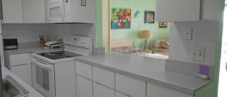 A kitchen with white appliances, a microwave, stove, and oven. There is a bed with a floral bedspread and a lamp.