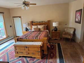 Une chambre à coucher avec un lit en bois, deux tables de chevet avec des lampes, une chaise, un tapis à motifs et un ventilateur de plafond.