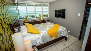 A bedroom with a bed, bedside lamps, a window with a view, a TV, and a small table with a lamp. - ENCHANTED Jewel @ Playa Azul Luquillo Beach 9th Floor (Luquillo)