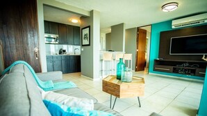 A living room with a grey sofa, a wooden coffee table, and a TV. There is a kitchen in the background with cabinets and a microwave. - ENCHANTED Jewel @ Playa Azul Luquillo Beach 9th Floor (Luquillo)