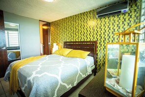 A hotel room with a bed, a bedside table, a lamp, a window, and a wall air conditioner.