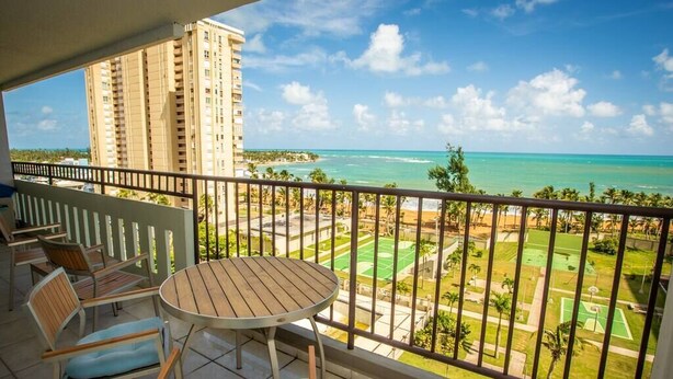 A balcony with a table and chairs, overlooking a beach and a sports court. - ENCHANTED Jewel @ Playa Azul Luquillo Beach 9th Floor (Luquillo)