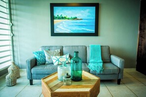 A living room with a grey sofa, a framed beach painting, and a coffee table with decorative items. - ENCHANTED Jewel @ Playa Azul Luquillo Beach 9th Floor (Luquillo)