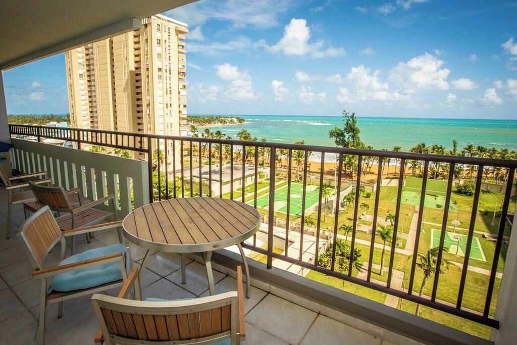 A balcony with a table and chairs, overlooking a beach and a sports court.