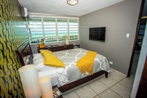 A bedroom with a bed, bedside lamps, a window with a view, a flat-screen TV, and a patterned wall.