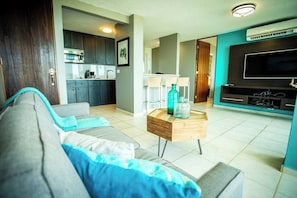 A living room with a sofa, a coffee table, and a TV. There is a kitchen in the background.