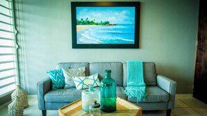 A living room with a grey sofa, a wooden coffee table, and a framed beach painting on the wall. - ENCHANTED Jewel @ Playa Azul Luquillo Beach 9th Floor (Luquillo)