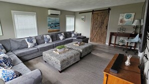 A living room with a grey sectional sofa, patterned ottoman, and a wooden coffee table. There are wall-mounted air conditioners and artwork on the walls.