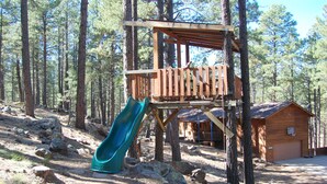 A wooden play structure with a slide in a forested area, surrounded by trees and a small cabin.