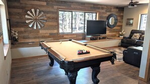 A pool table with a billiard ball rack, a television, and a leather sofa in a room with wooden flooring and a large window.