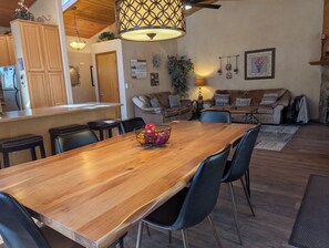 A wooden dining table with chairs, a fruit bowl, a kitchen area with cabinets, and a living room with a sofa and decorative items.
