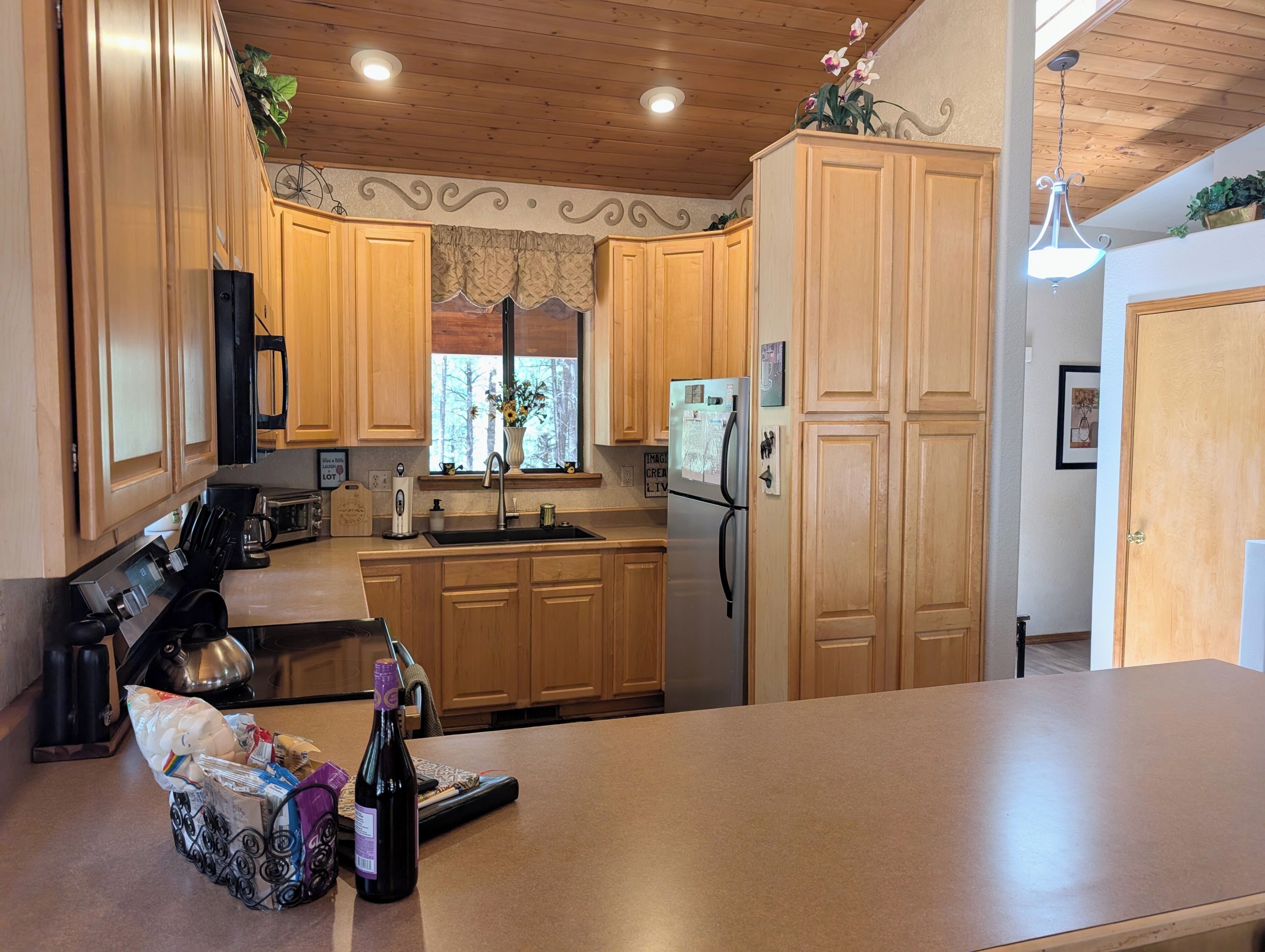 A kitchen with wooden cabinets, a stainless steel refrigerator, a sink, and a window with a view of trees.