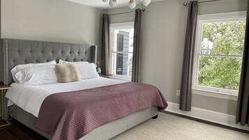 Una camera da letto con una testiera trapuntata, un letto grande con biancheria da letto bianco e beige, una coperta trapuntata viola e vista sugli alberi attraverso una finestra.