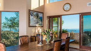 A dining area with a wooden table, leather chairs, a vase with flowers, a clock on the wall, and a scenic view through large windows. - Private Vineyard Estate Overlooking Pacific Ocean (Malibu)