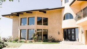 A modern two-story house with large windows, a curved roof, and a well-maintained garden. - Private Vineyard Estate Overlooking Pacific Ocean (Malibu)