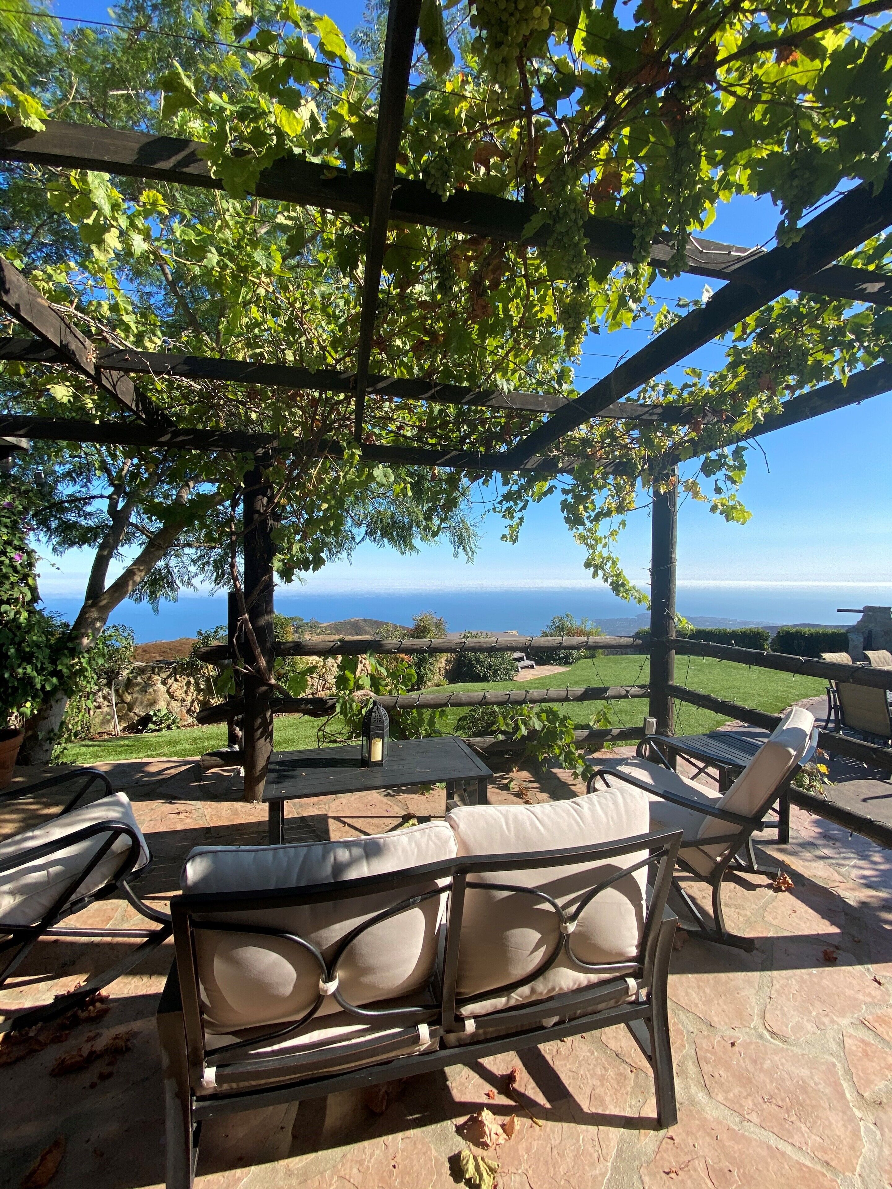 An outdoor seating area with a pergola, tables, and chairs, overlooking a scenic landscape with a view of the sea.