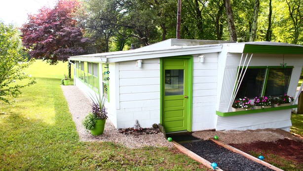 Une petite maison de plain-pied avec une porte et des fenêtres vertes, entourée d’une pelouse bien entretenue et d’arbres.