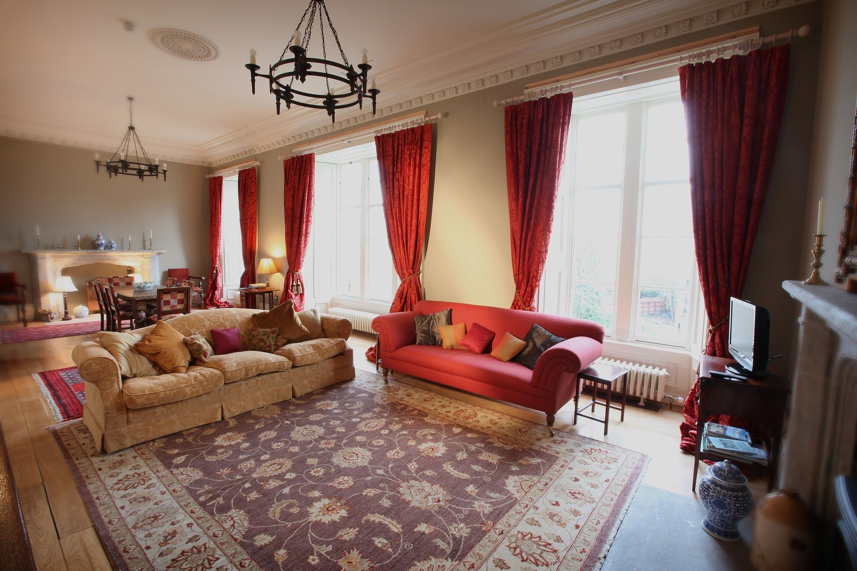 Amplia sala de estar con un sofá rojo grande, un sofá color beige y un tapete estampado. Hay una chimenea con repisa, un candelabro y un televisor sobre un mueble.