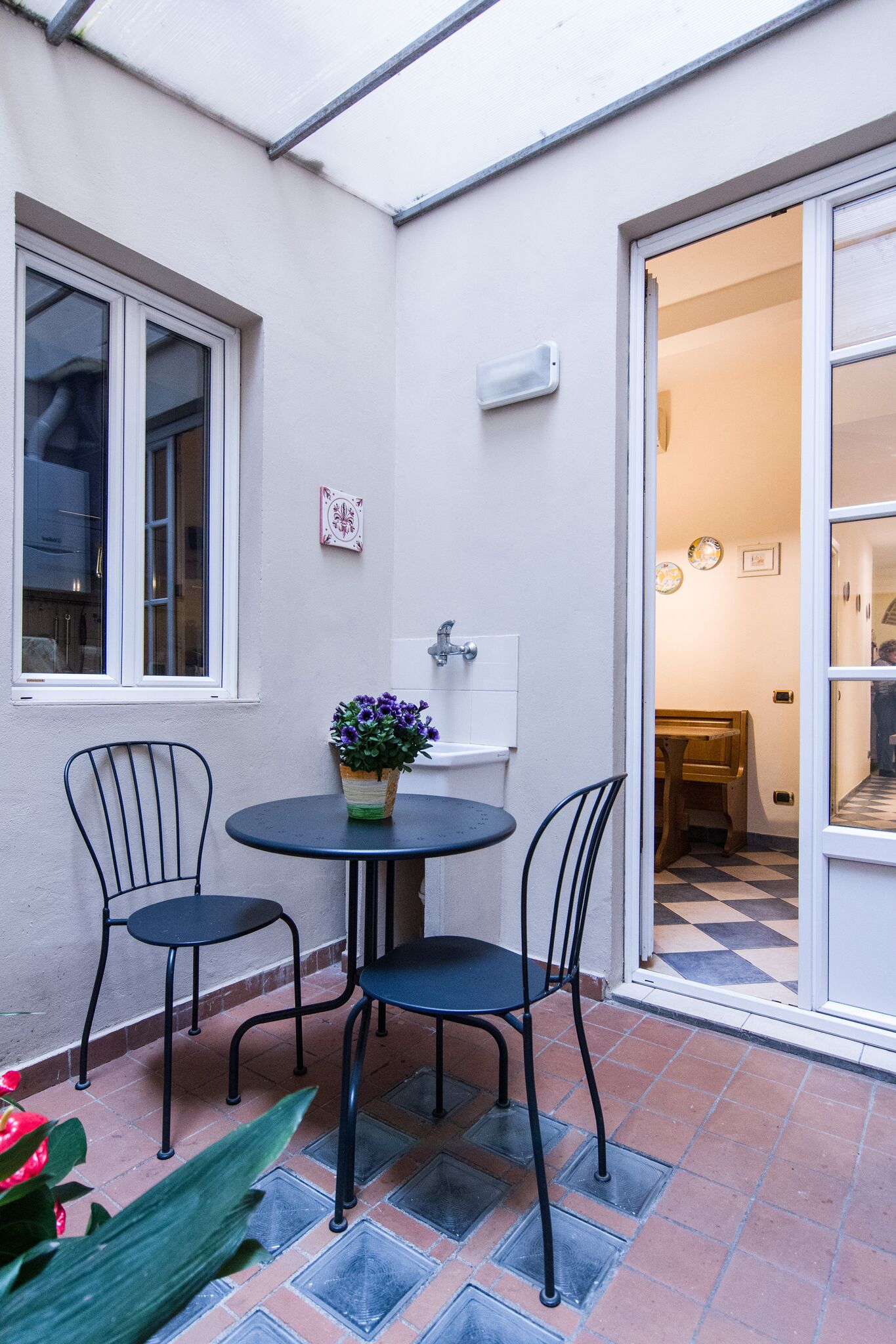 Eine kleine Terrasse mit einem runden Tisch, zwei Stühlen, einem Blumentopf und einem Fenster.