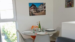 Un comedor con una mesa lista para dos personas, un sofá con un cojín colorido y una ventana con vista a follaje.