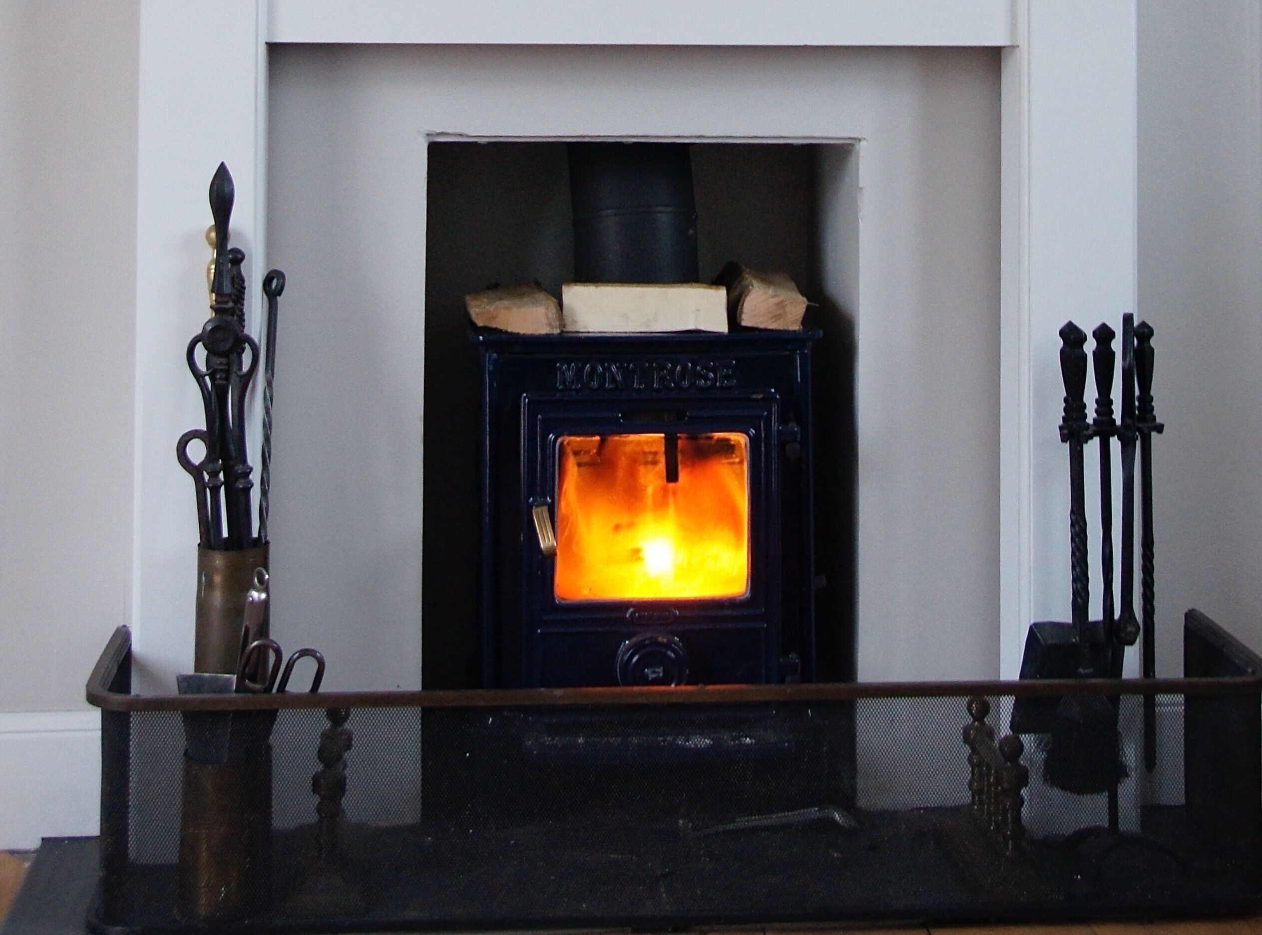 A traditional wood-burning stove with a visible fire, set in a white-walled room with a metal grate in front.