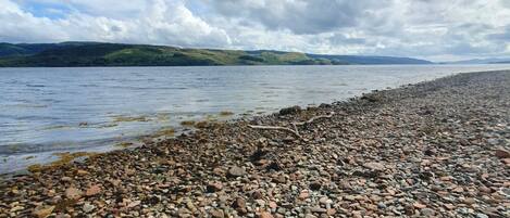 A pebbled beach with a view of a lake and distant hills under a cloudy sky.