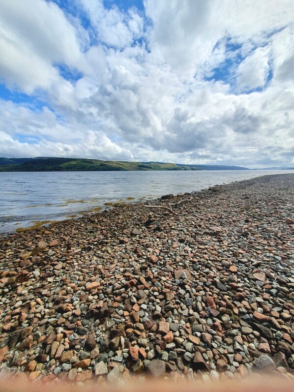 A pebbled beach with a view of a lake and distant hills under a cloudy sky.
