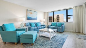 Un salón moderno con muebles color turquesa, un ventanal con vistas a edificios y palmeras, y un cuadro en la pared.