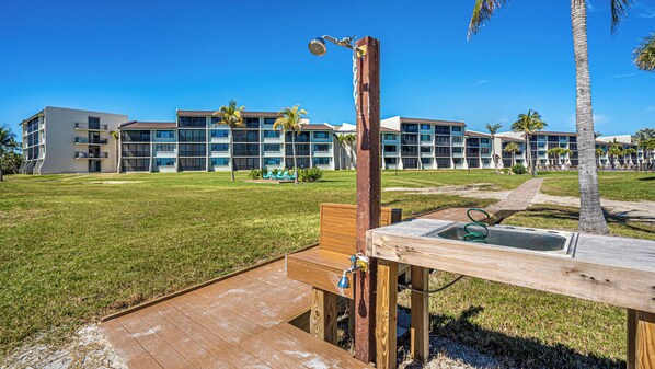 A wooden deck with a sink and shower, surrounded by a grassy area and palm trees, with modern buildings in the background. - FABULOUS GULF FRONT, 2/2 condo, PET FRIENDLY, 4 bikes, Pool, pickleball, tennis! (Sanibel)