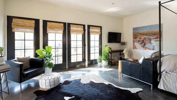 A modern living room with a black leather sofa, a black and white cowhide rug, and large windows with bamboo blinds.