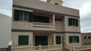 A two-story building with green shutters, a balcony with a patterned railing, and a stone staircase leading to the entrance.