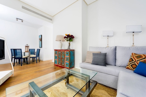 A modern living room with a glass coffee table, a grey sofa, and a dining area with blue chairs.