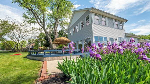 A two-story house with a large garden, a thatched umbrella, and purple flowers. - 4 Bedroom Waterview - MD Chesapeake Bay! Walk to Marina, Beach Bar & Restaurant! (Stevensville)