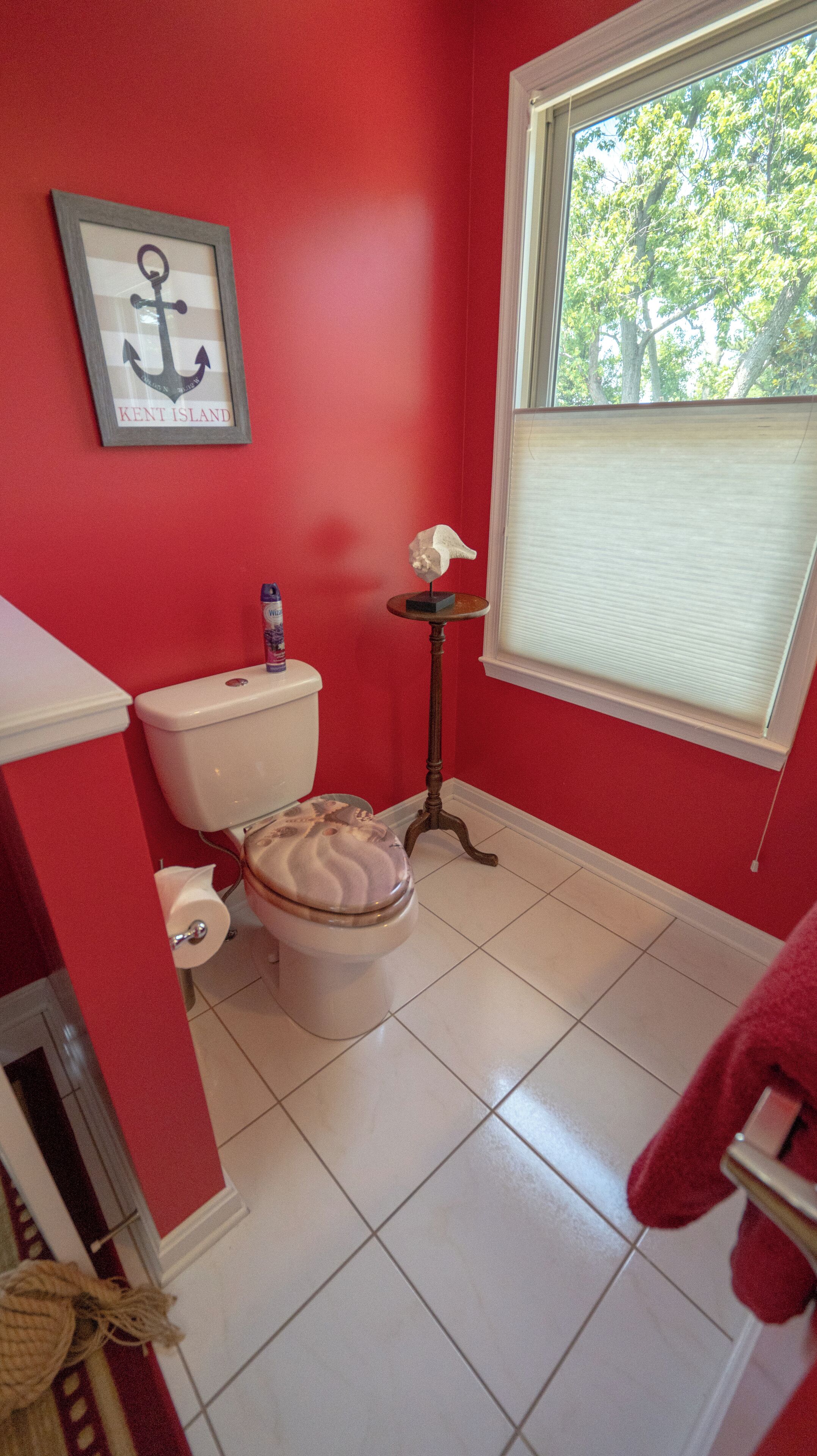 A bathroom with a red wall, a toilet, a framed anchor picture, a small table with a tissue box, and a window with blinds.