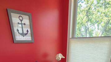 A bathroom with a red wall, a toilet, a framed anchor picture, a small table with a tissue box, and a window with blinds.