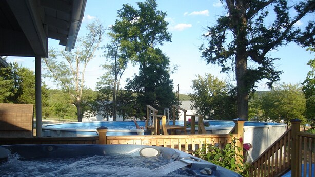 A hot tub area with wooden decks and a view of trees and a building in the background. - Quiet, Private, 2 acres to enjoy and still close to everywhere you want to go. (Elkins)