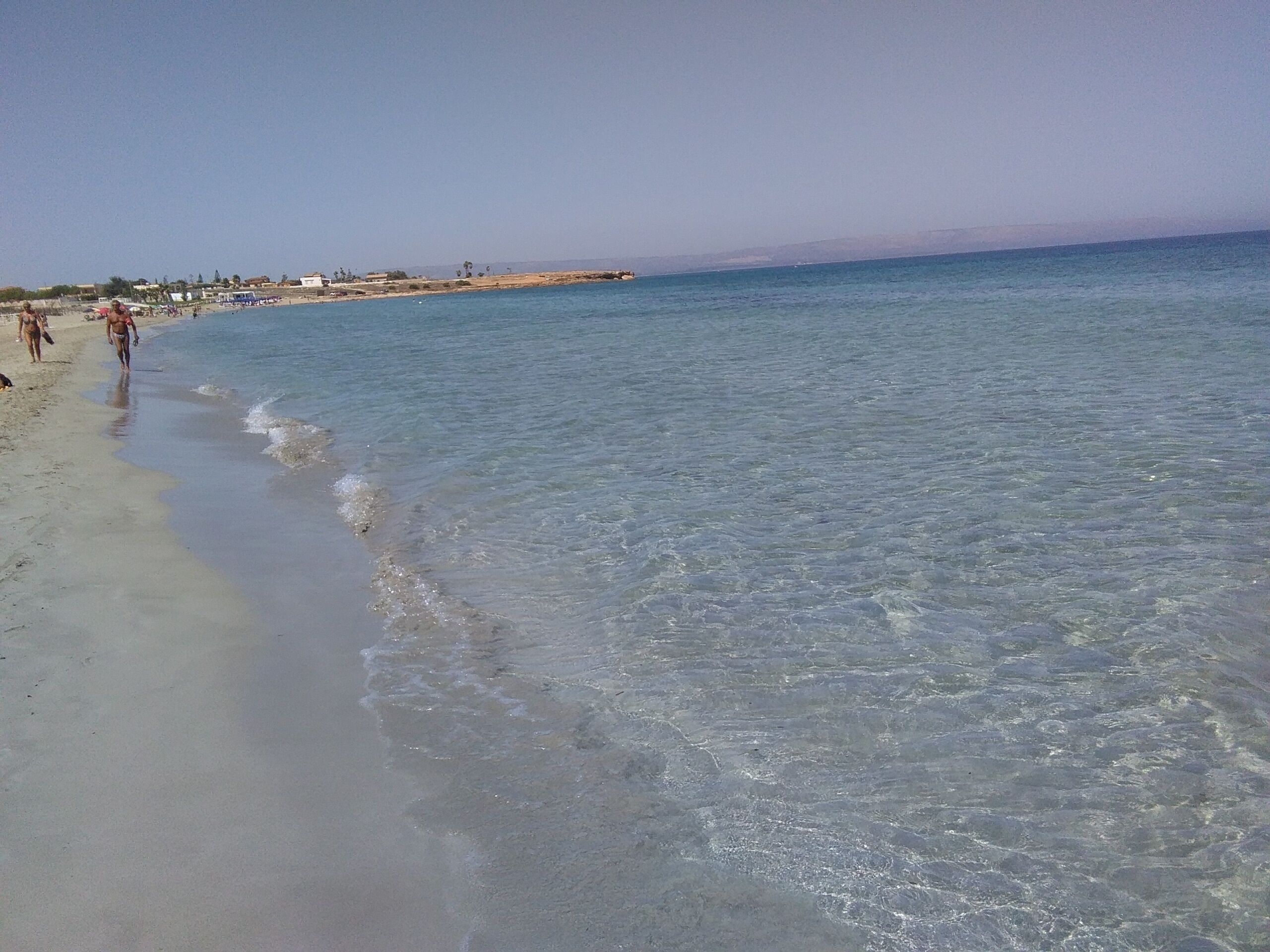 En strand med klart, lavt vand, en sandet kystlinje og et par personer, der går en tur.
