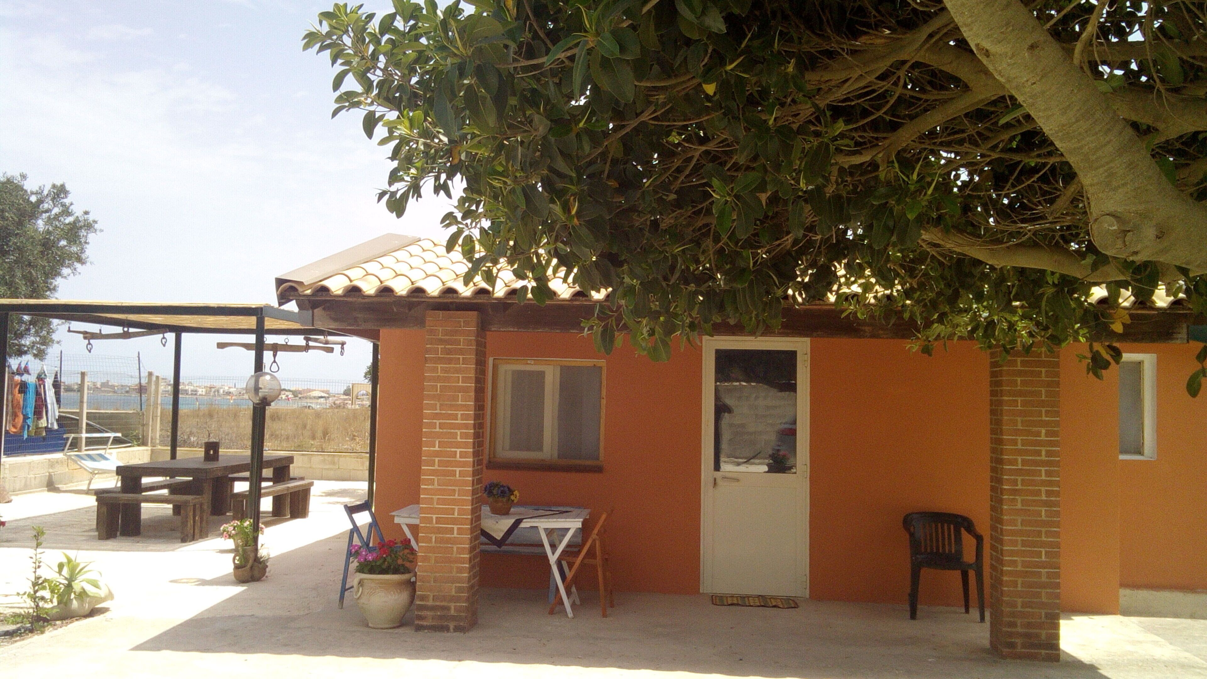 Un pequeño edificio de ladrillo con techo de tejas, una puerta y una ventana. Hay una mesa y una silla afuera, y un árbol proporciona sombra.