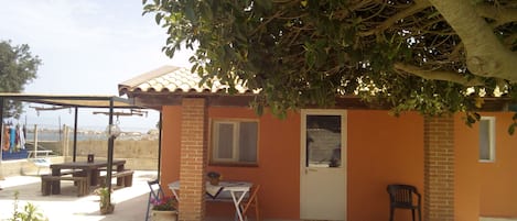 A small brick building with a tiled roof, a door, and a window. There is a table and a chair outside, and a tree provides shade.