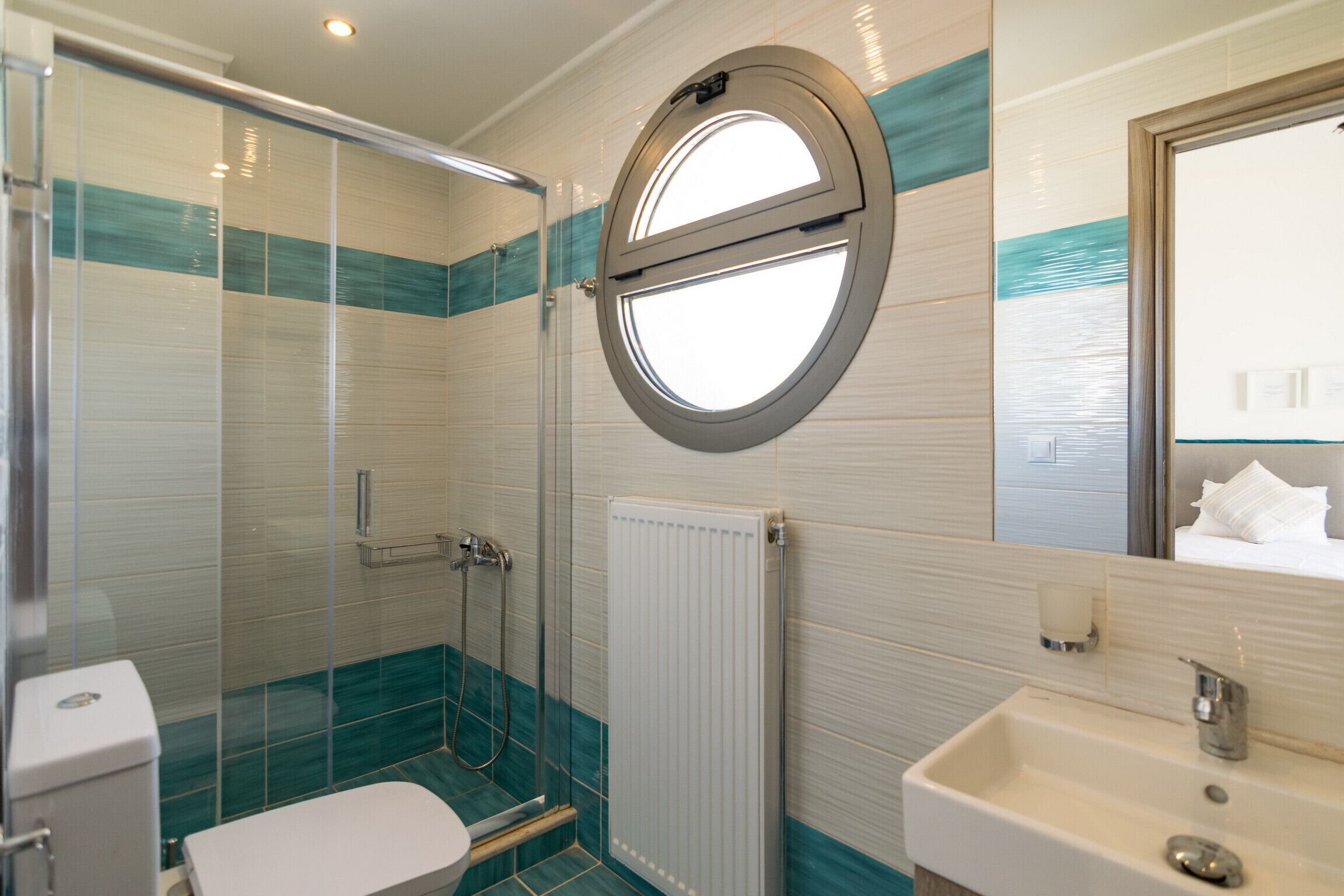 A bathroom with a glass shower enclosure, a toilet, a sink, and a round mirror with a window.