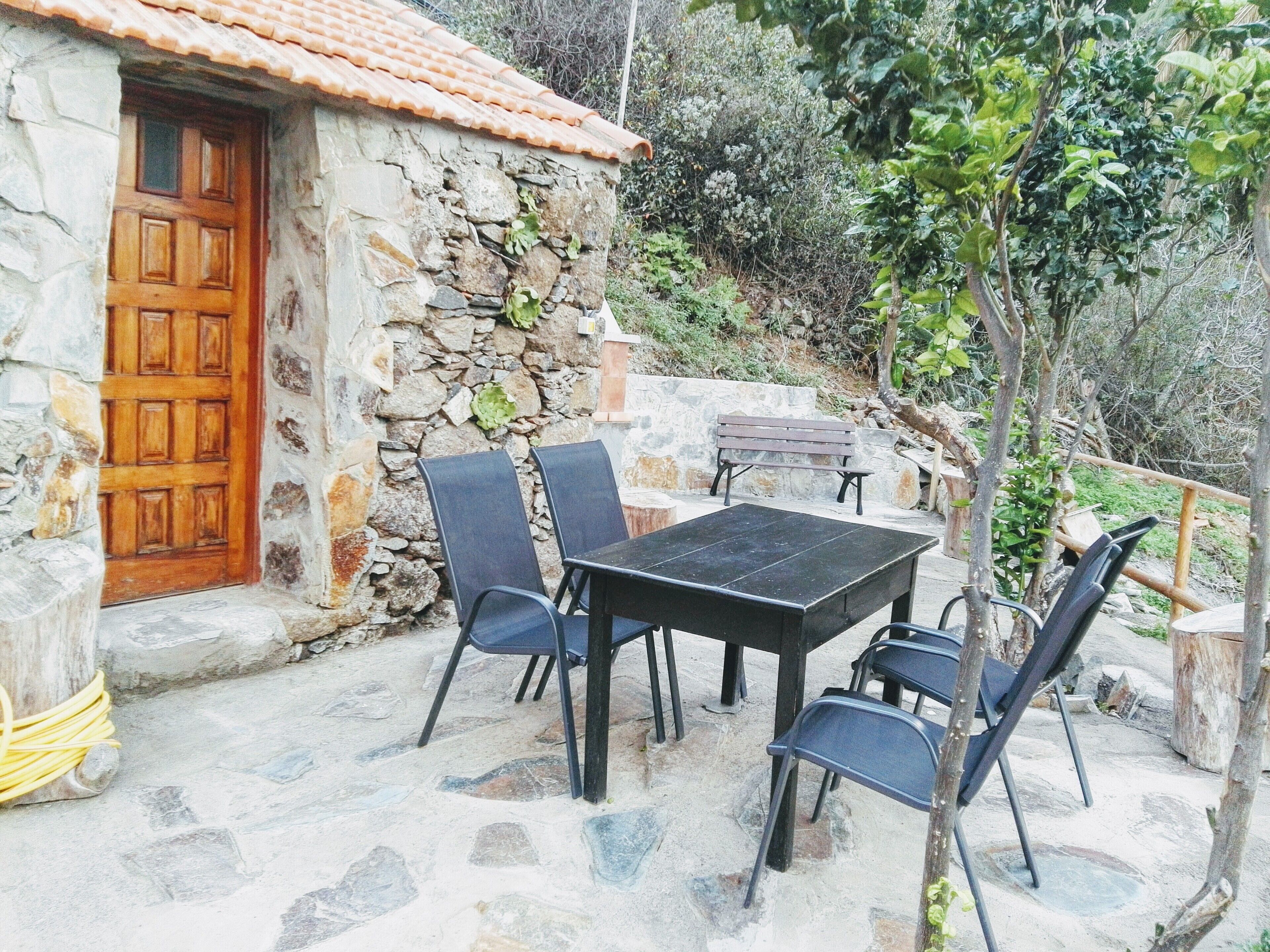 Un edificio de piedra con puerta de madera, pared de piedra con plantas y un patio con mesa y sillas.