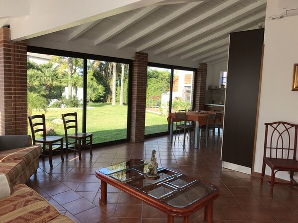 A spacious living area with a glass door leading to a garden, a dining table with chairs, and a sofa.