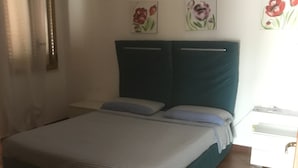 Ein Schlafzimmer mit einem Bett, zwei Kissen, einem Nachttisch und einer Kommode. Darüber befinden sich Wandbilder mit floralem Muster.