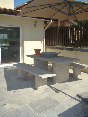 Een terras buiten met een stenen tafel en bankjes, een potplant en een glazen schuifdeur naar een keuken.